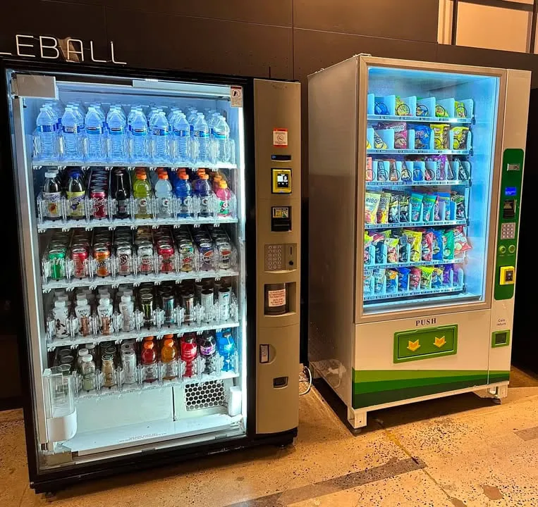 Glass-front cold drink vending machine stocked with bottled water, sports drinks and sodas installed beside a glass-front snack machine in an office lobby