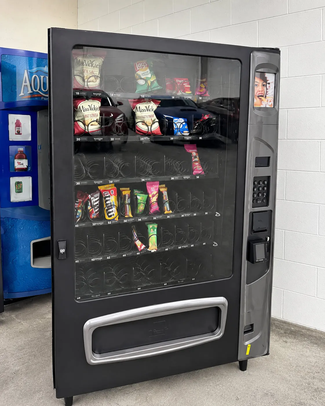 Glass-front snack vending machine with a credit card and tap-to-pay reader, stocked with chips, candy and cookies beside a beverage machine
