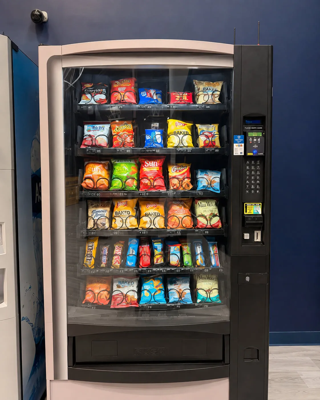 Modern glass-front snack vending machine stocked with chips, baked snacks and candy bars in a corporate lobby, with keypad and card reader